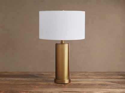 Linden Table Lamp with a brass-finished cylindrical base and a glowing white linen drum shade on a rustic wooden table.