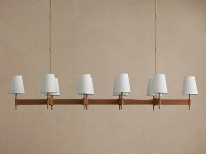 Hastings Linear Chandelier with brass structure, brown leather bar, six lit white linen shades, against a beige wall.
