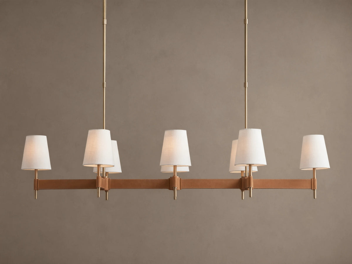 Hastings Linear Chandelier with 6 white linen shades, antiqued brass frame, and brown leather details on a plain wall.