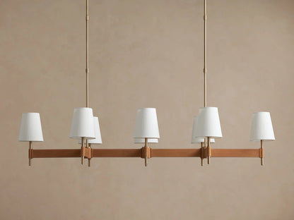 Hastings Linear Chandelier with 6 white linen shades, antiqued brass frame, and brown leather details on a plain wall.