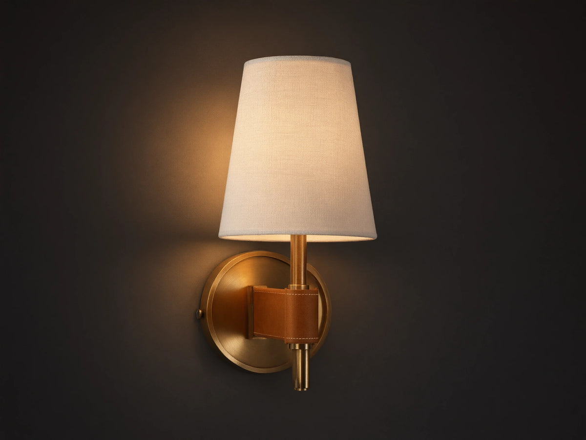 Hastings Sconce: Lit, antiqued brass wall sconce with a tapered white linen shade and brown leather accent, on a neutral wall.