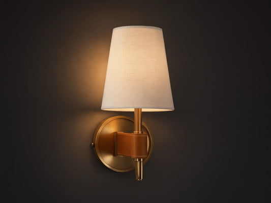 Hastings Sconce: Lit, antiqued brass wall sconce with a tapered white linen shade and brown leather accent, on a neutral wall.