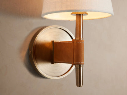 Hastings Sconce on a beige wall. It has an antiqued brass base, brown leather detail, and a white tapered linen shade.