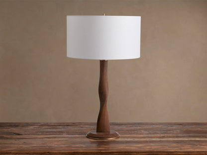 Jonas Table Lamp: White linen shade, curved dark oak wood base with brass accents, on a rustic wooden table.