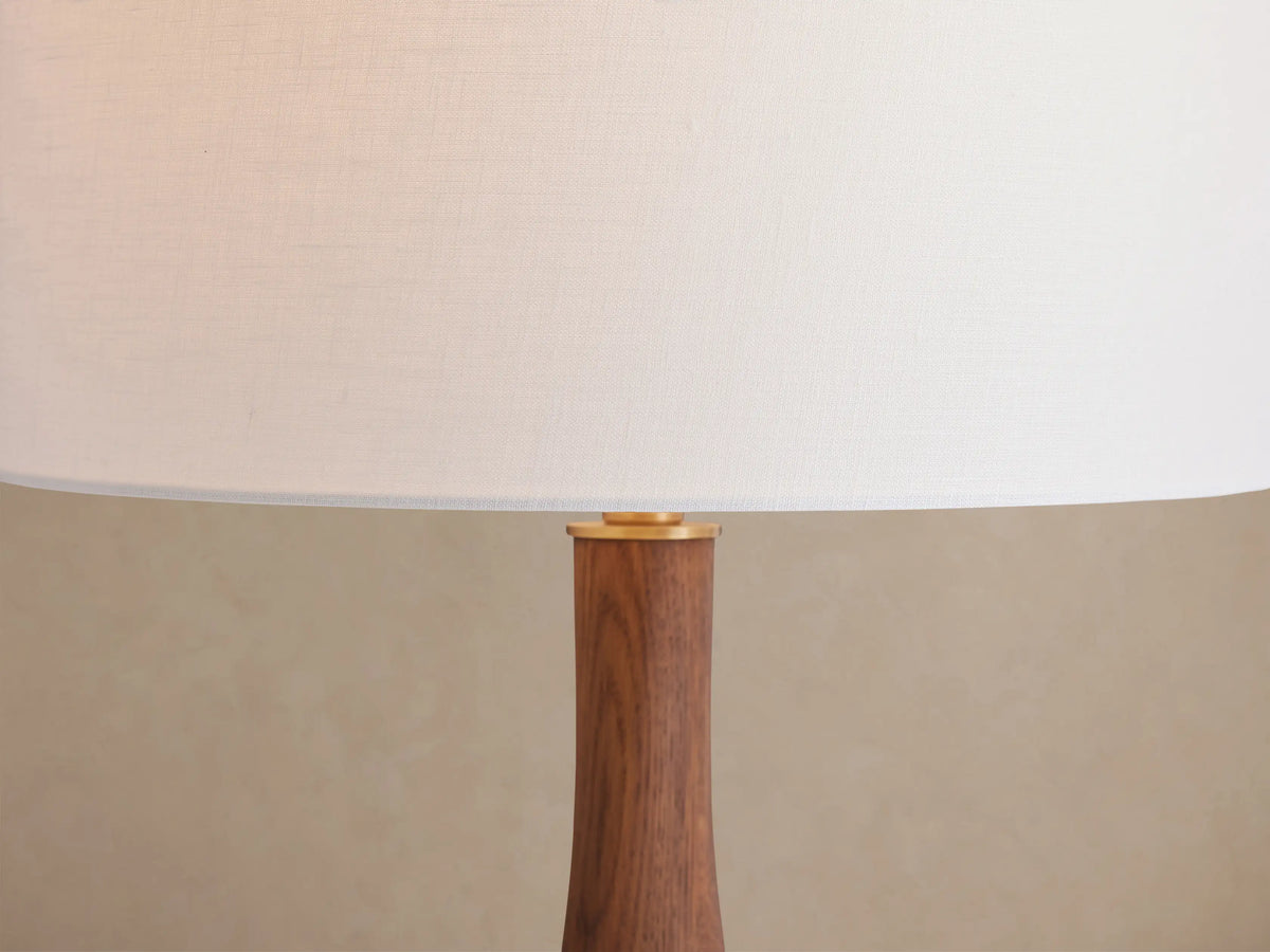 Jonas Table Lamp with white linen shade, curved dark wood stem, and brass accents, on a rustic wood table.
