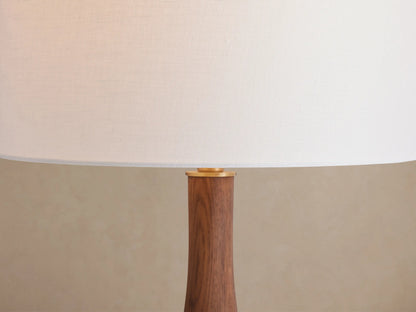 Jonas Table Lamp with white linen shade, curved dark wood stem, and brass accents, on a rustic wood table.