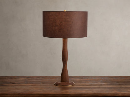 A brown linen shade tops the Jonas Table Lamp, crafted from oak wood with a twisted design and brass accents, on a wooden table.