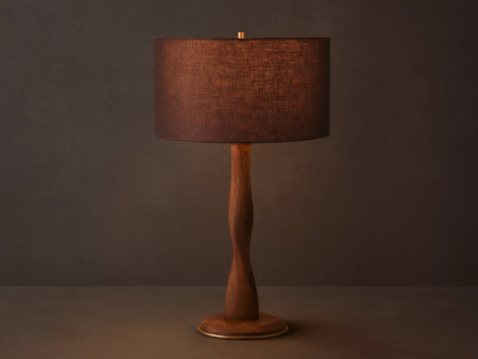 A brown linen shade tops the Jonas Table Lamp, crafted from oak wood with a twisted design and brass accents, on a wooden table.