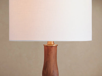 Jonas Table Lamp with a white linen shade and a twisted dark oak wood base with brass accents, on a rustic wooden table.