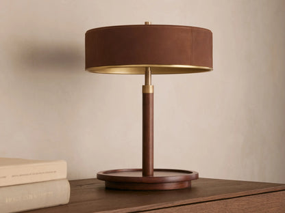 A wooden table lamp with a round dark brown shade, emitting warm light, sits on a wooden surface beside closed books.