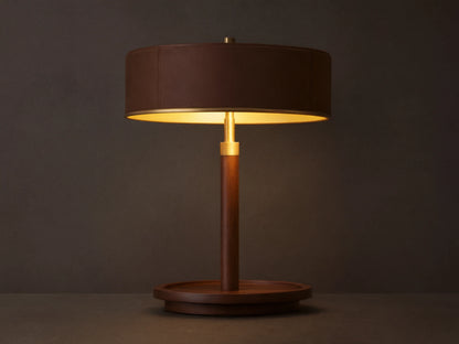A wooden table lamp with a round dark brown shade, emitting warm light, sits on a wooden surface beside closed books.