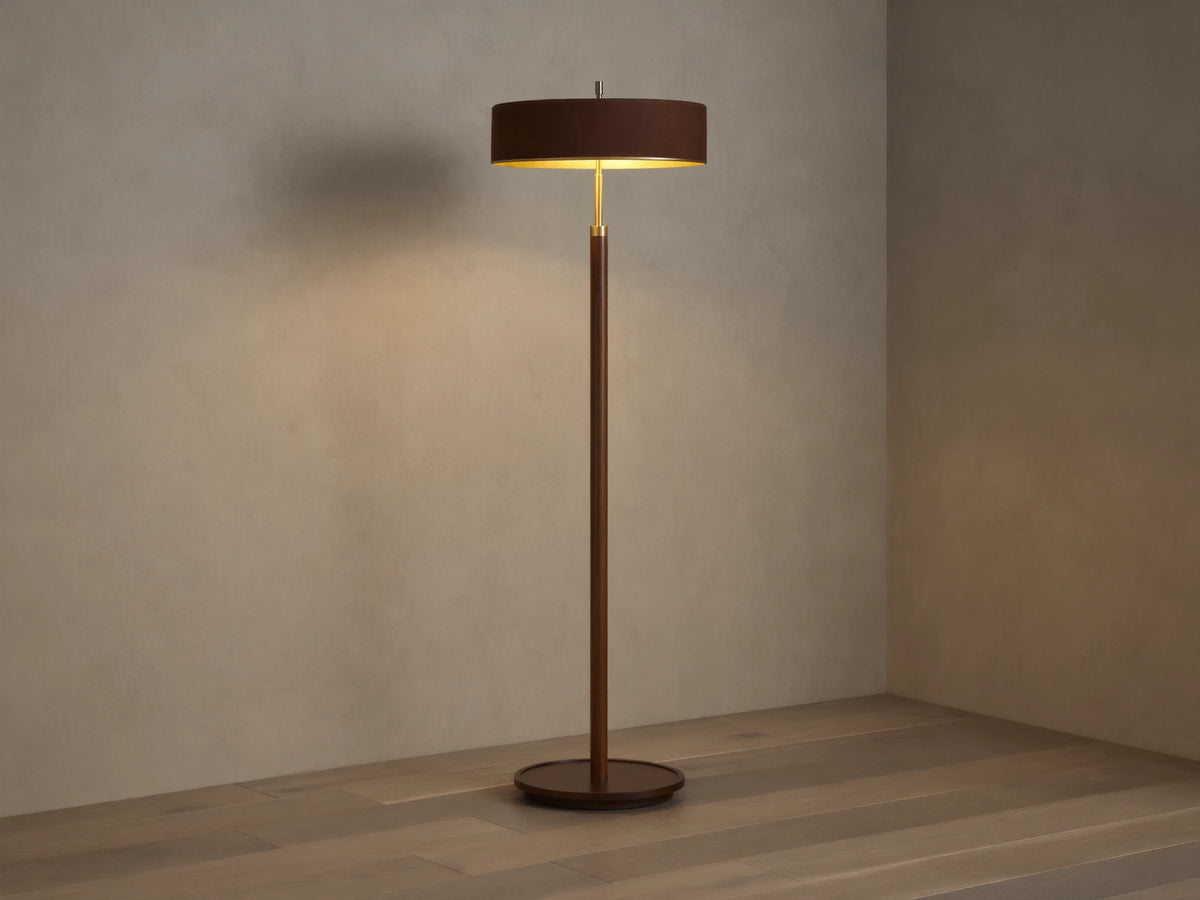 A modern floor lamp with a brown cylindrical shade and slender wooden stand, casting warm light on a wooden floor and neutral wall.