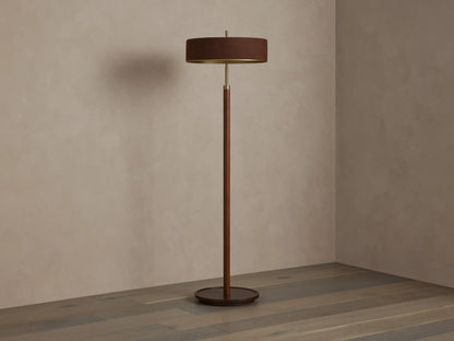 A modern floor lamp with a brown cylindrical shade and slender wooden stand, casting warm light on a wooden floor and neutral wall.