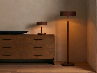 A modern lamp with a wide, dark brown drum shade and gold accents, warmly lit on a textured neutral background.