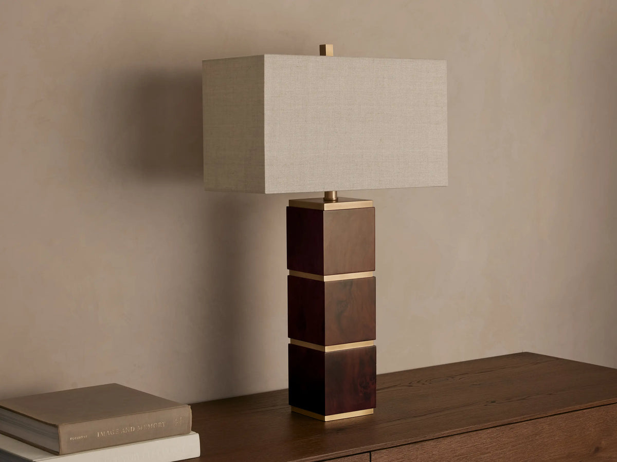 A table lamp with a rectangular fabric shade and a base composed of stacked wooden cubes with gold accents, on a wooden surface.