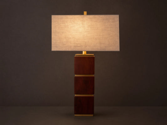 A table lamp with a rectangular fabric shade and a base composed of stacked wooden cubes with gold accents, on a wooden surface.