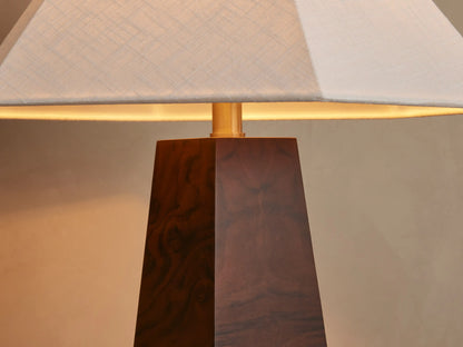 A table lamp with a wooden pyramid-shaped base and a wide, white, tapered square lampshade, placed on a wooden surface next to books.