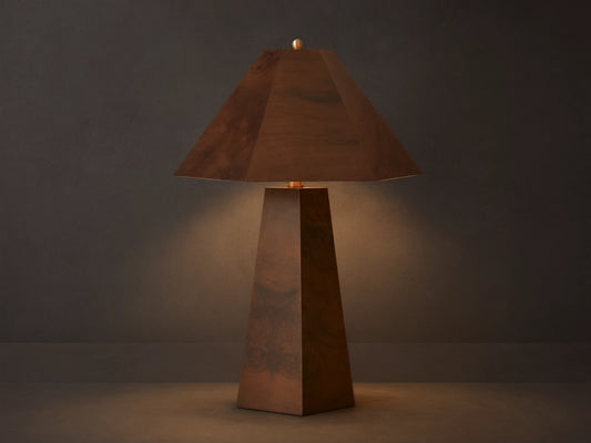 A wooden table lamp with a pyramid-shaped shade and tapered base emits a warm glow on a wooden surface beside stacked books.