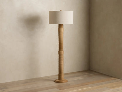 Tall light wood floor lamp with a ribbed stand and glowing beige fabric drum shade, standing in a room corner.