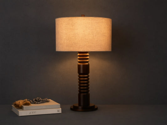 Front view of a dark brown carved base table lamp with a cylindrical light beige fabric shade, illuminated.