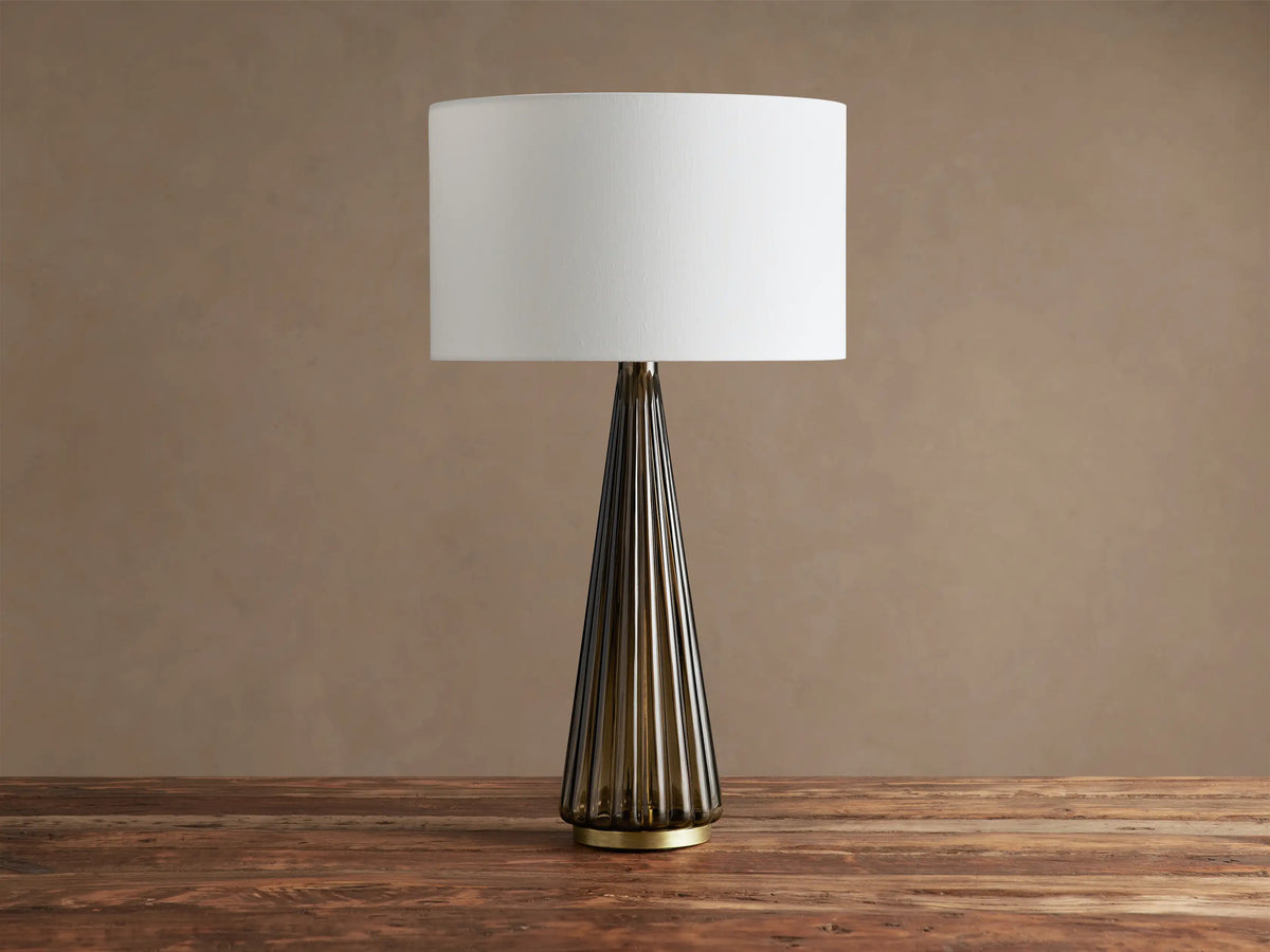 Elisabetta Table Lamp: upright, white linen shade, smoky fluted glass base, brass accent, sitting on a wooden table.