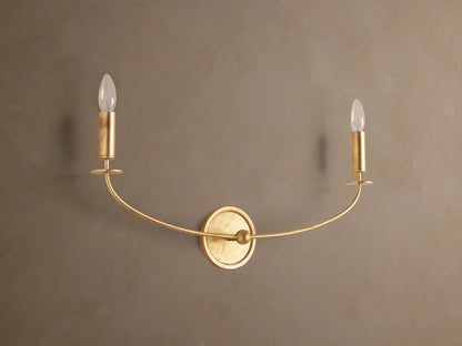 Arnett Double Sconce with an antiqued gold finish, featuring a round backplate and two candle-style lights on a curved arm.