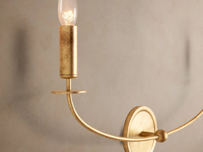 Arnett Double Sconce: antiqued gold iron wall light with two curved arms holding candlestick-style clear bulbs on a textured wall.