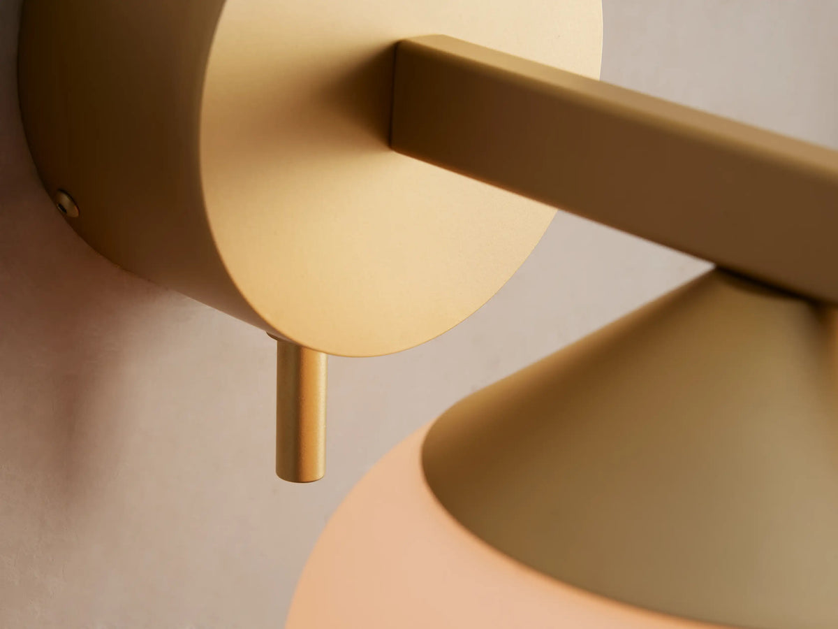 Costella Sconce with a gold circular backplate and arm supporting a conical gold piece and a round white opal glass globe diffuser on a taupe wall.