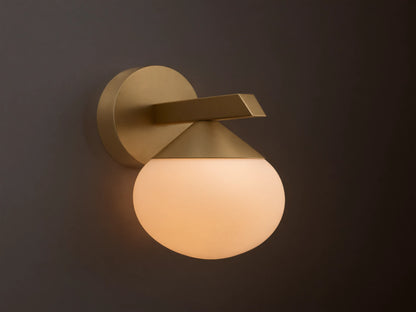 Costella Sconce, a gold iron wall light with a geometric arm and an illuminated, round opal glass diffuser, mounted on a neutral wall.