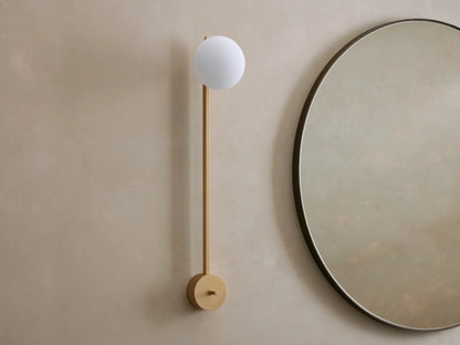 Costella Grand Sconce: a long champagne gold iron wall light with a glowing white spherical opal diffuser, next to a round mirror.