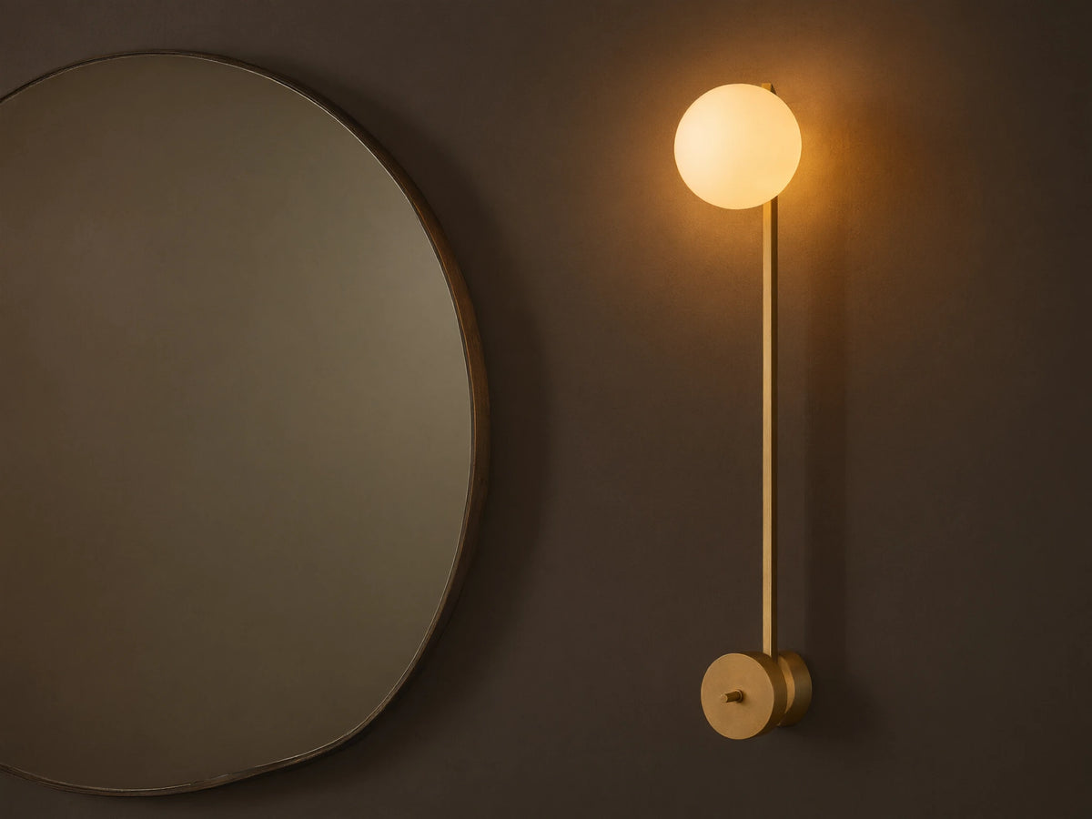 Costella Grand Sconce: a long champagne gold iron wall light with a glowing white spherical opal diffuser, next to a round mirror.