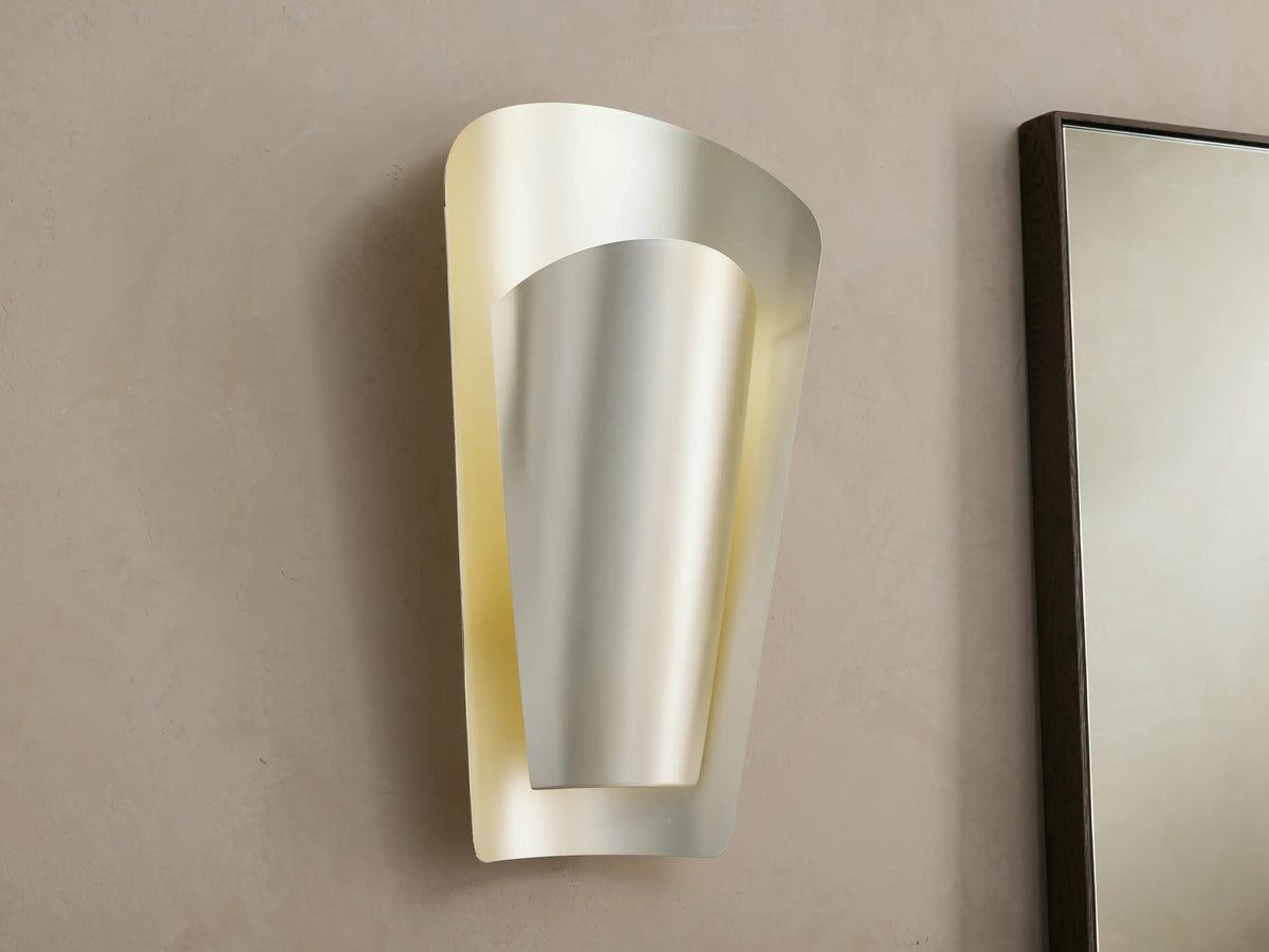 Illuminated Strato Sconce on a textured wall. Features a metallic leafed finish, curved outer plate, and inner diffuser emitting warm light.