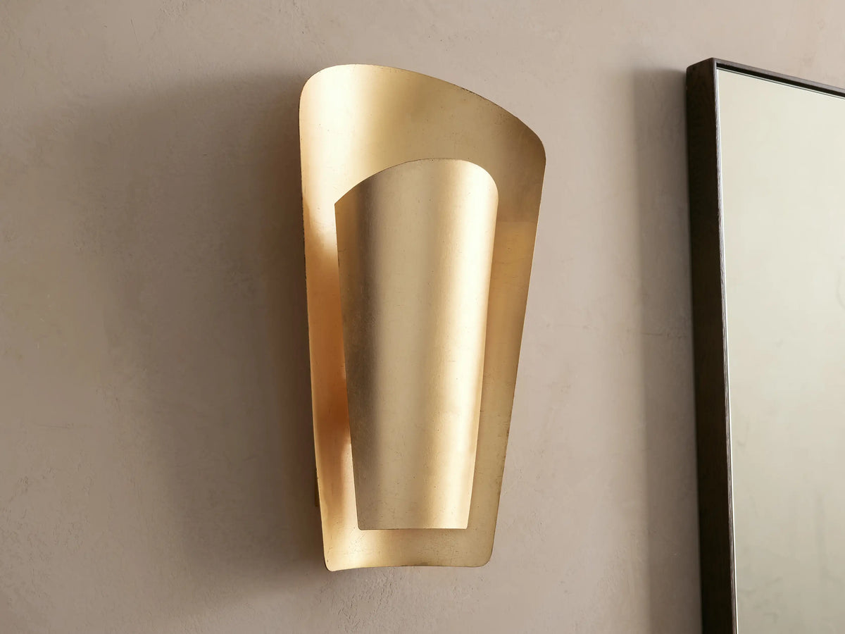 Strato Sconce, a curved, vertical, gold-leafed brass wall sconce with a modern design, illuminated against a neutral wall.