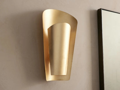Strato Sconce, a curved, vertical, gold-leafed brass wall sconce with a modern design, illuminated against a neutral wall.