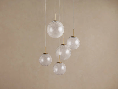 Casen Round Chandelier with five clear crackle glass globes, each suspended by a thin wire and a satin brass stem, against a beige wall.