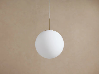 Casen Pendant: Illuminated white opal glass globe suspended from a satin brass rod and thin wire against a warm wall.