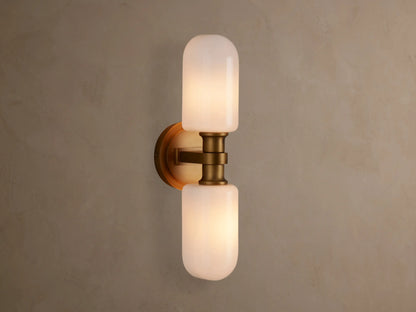 Zayd Double Sconce, vertically mounted, with two illuminated white cylindrical glass shades and brass-toned hardware on a textured wall.