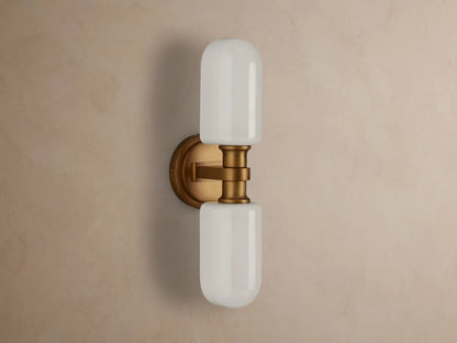 Zayd Double Sconce, vertically mounted, with two illuminated white cylindrical glass shades and brass-toned hardware on a textured wall.