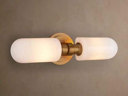 Zayd Double Sconce: Vertical brushed brass fixture with two illuminated frosted white glass shades, on a gray wall.