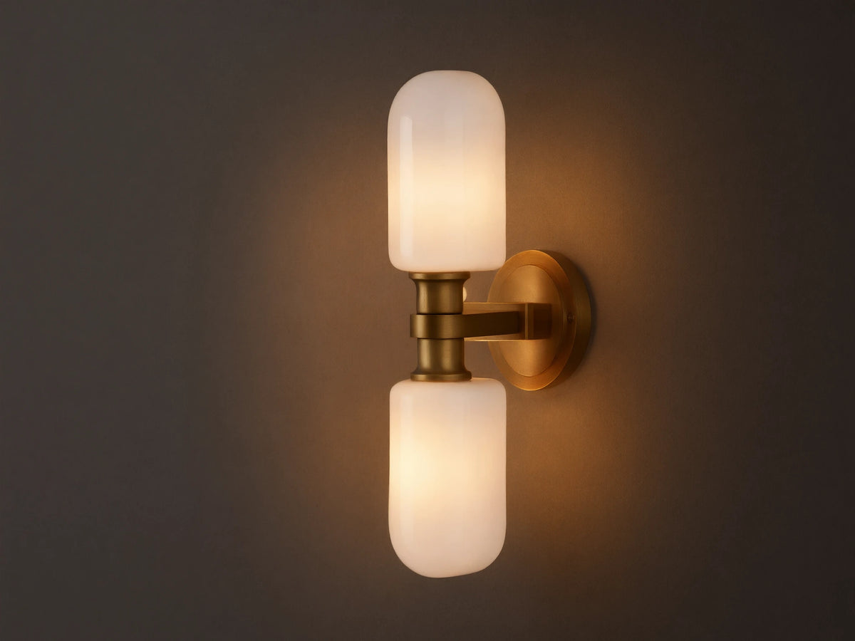 Zayd Double Sconce, vertically mounted, with two illuminated white cylindrical glass shades and brass-toned hardware on a textured wall.