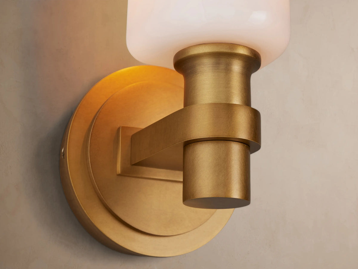 Zayd Sconce: Wall-mounted brass light with a vertical, rounded white opal glass shade, on a textured beige wall.