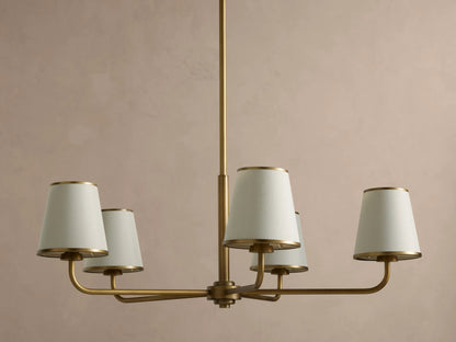 Adria Round Chandelier: brass fixture with 5 cream conical shades, gold metal trim, hanging from a rod on a beige wall.