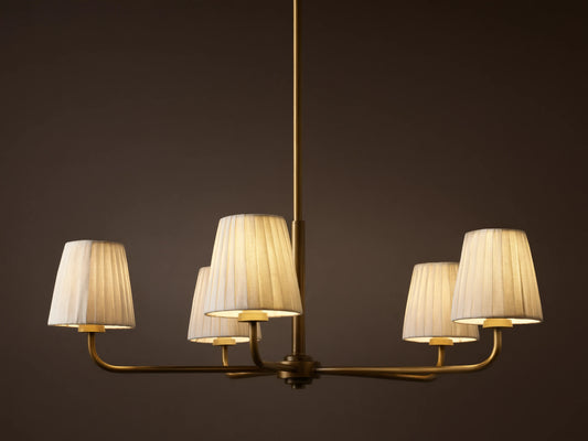 Adria Round Chandelier with five illuminated pleated white shades on a brass-finished frame, hanging against a dark wall.
