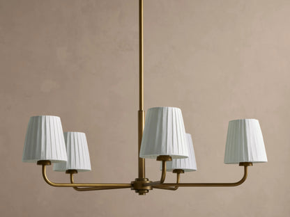 Adria Round Chandelier: golden iron frame with five white pleated shades, hanging against a light beige wall.