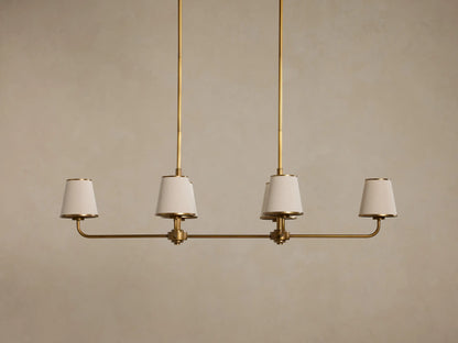 Adria Linear Chandelier with four glowing, tapered beige fabric shades trimmed in brass, suspended by two brass rods.