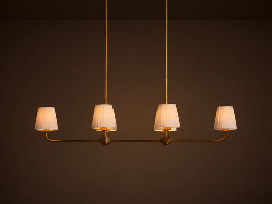 Adria Linear Chandelier with 4 illuminated pleated white shades on a horizontal brass bar, hanging from 2 brass rods.