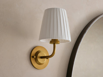 Adria Sconce: brass fixture, pleated beige shade, warm light on a textured wall beside a dark wood mirror.