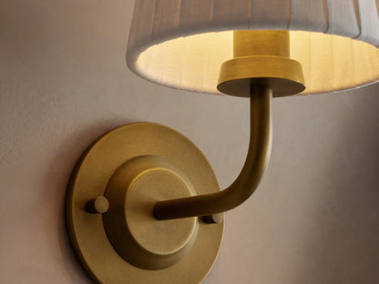 Adria Sconce featuring a round brass base, curved brass arm, and a white pleated fabric shade, mounted on a light wall.