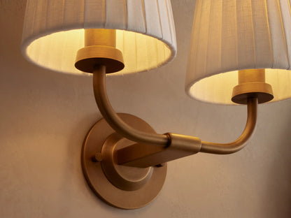 Adria Double Sconce: Two white pleated shades on a curved brass-finished iron frame, mounted on a beige wall. Partial mirror right.