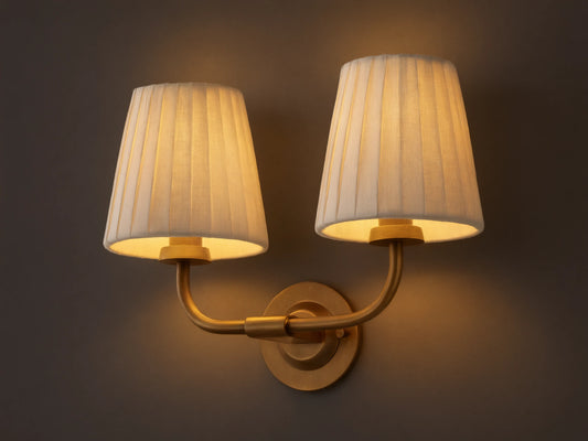 Adria Double Sconce with two lit off-white pleated shades on a brass-toned iron base, mounted on a wall next to a mirror.